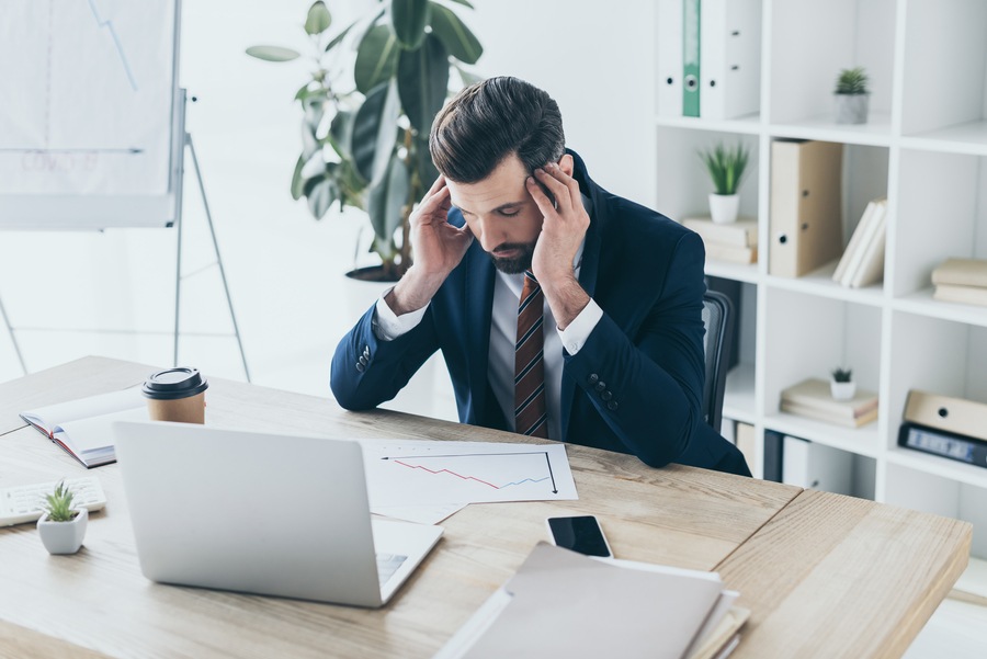 depressed-businessman-touching bowed head while sitting at workplace with closed eyes